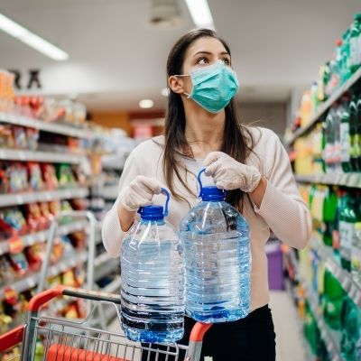 Eine Frau mit Gesichtsmaske und Handschuhen steht in einem Lebensmittelgeschäft und hält zwei große Wasserflaschen über einem Einkaufswagen, umgeben von Regalen mit verschiedenen Produkten.