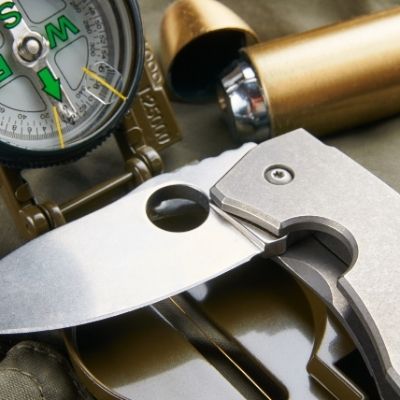 Eine Nahaufnahme eines Klappmessers aus Metall, eines Kompasses mit grünen Markierungen und eines zylindrischen Behälters aus Messing auf einem khakifarbenen Stoffhintergrund, was auf ein Outdoor- oder Survival-Thema hindeutet.